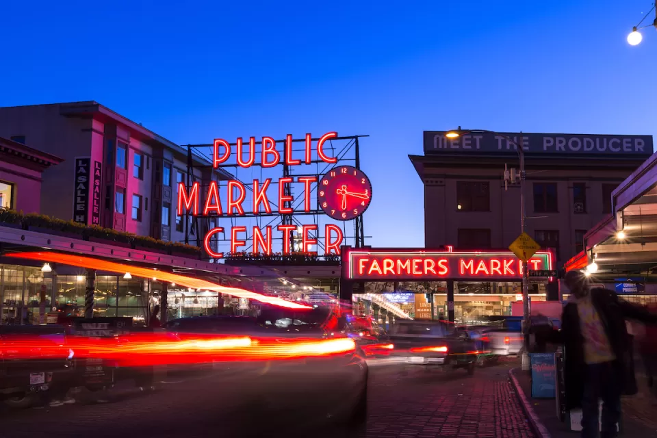 Pike Place Market in Seattle at twilight with neon market sign and cars passing through.
